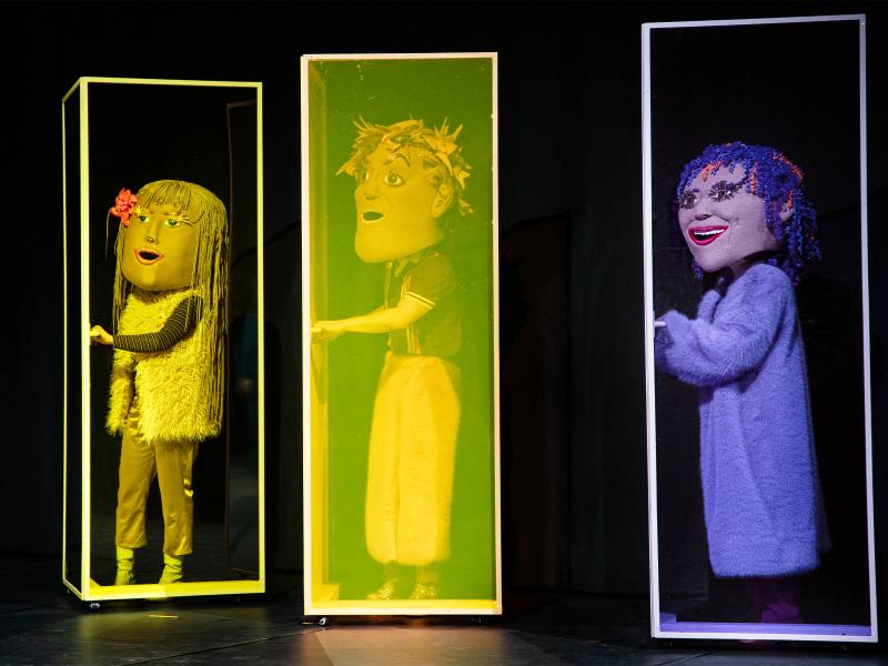 Bühnensituation: Drei Personen mit großen Kopfmasken stehen einzeln in drei hohen, eckigen Kästen. Alle haben die Hand auf einer Türklinke liegen.