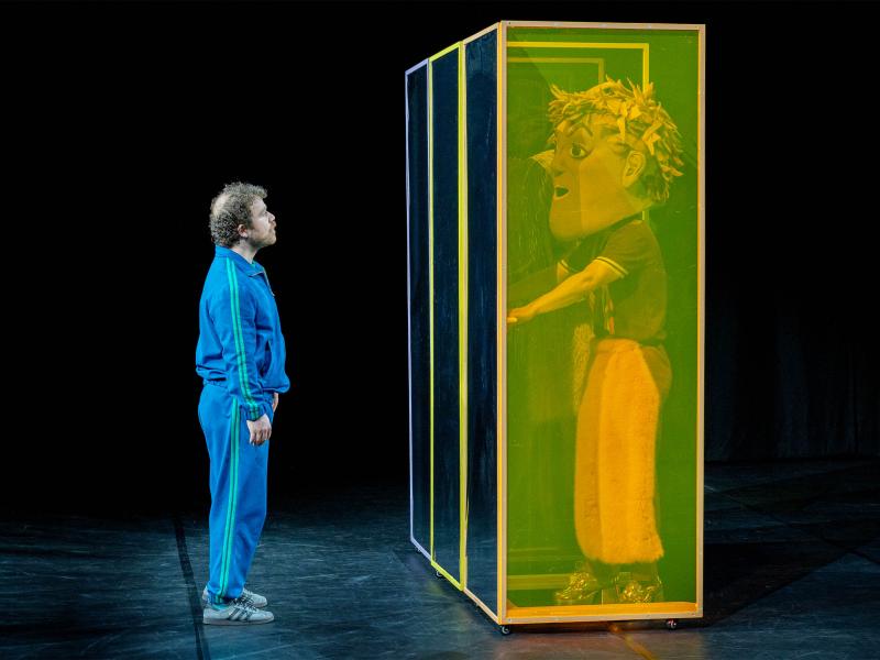 Bühnensituation: Parsa Yaghoubi Pour in einem blauen Trainingsanzug, steht vor drei nebeneinanderstehenden hohen eckigen Kästen. Die Seitenwand ist grünlich transparent. In einem Kasten steht eine Person in braun-rosafarbener Kleidung mit einer großen Kopfmaske mit braun-rosa Haaren mit der Hand an einer Türklinke. Parsa schaut sie an. 