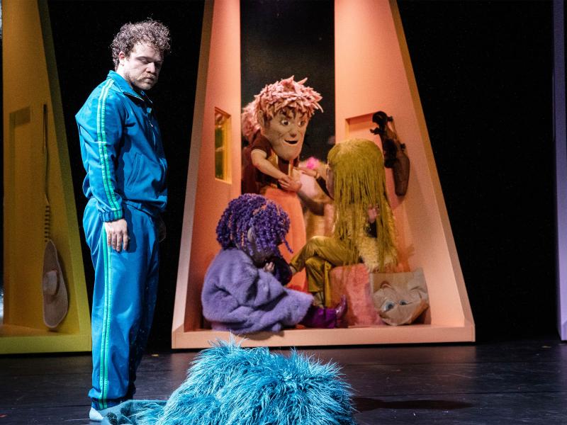 flauschiges blaues Stoffstück auf dem Boden. Im Hintergrund ein kleiner, dreieckiger hoher orangefarbener Mini-Raum, in dem sich drei Personen mit großen Kopfmasken befinden.