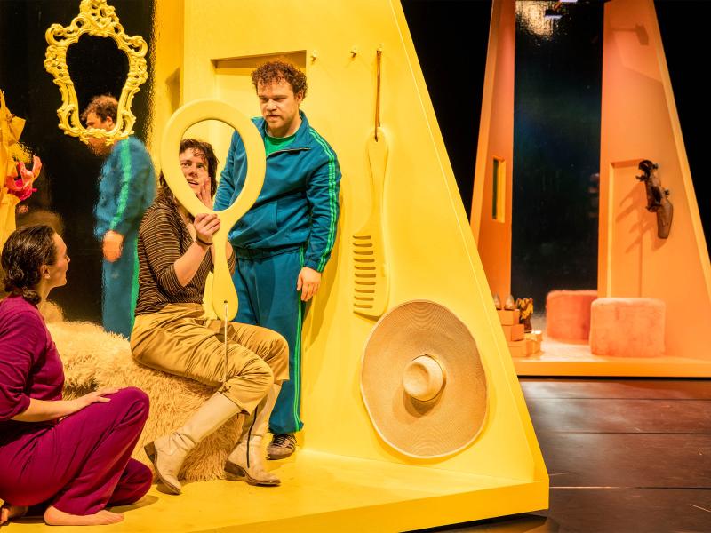 Bühnensituation: Das Ensemble trägt bunte, einfarbige Kleidung in rot, gold, blau oder rosa. Viktoria Kraft, Anastasia Lara Heller und Parsa Yaghoubi Pour befinden sich in einem gelben kleinen Raum. Ein übergroßer gelber Kamm und ein großer Sonnenhut hängen an der Wand, Anastasia schaut in einen übergroßen gelben Handspiegel ohne Spiegelfolie, eine Hand an die Wange gelegt. Im Hintergrund eine Person mit braun-rosa Kleidung und einer großen Kopfmaske mit Gesicht.