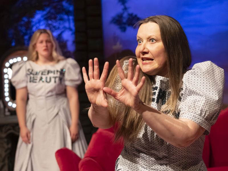 Bühnensituation: Christine Ochsenhofer und Anouk Piwek, beide in einem hellen gepunkteten Kleid mit Puffärmeln, auf ihrer Brust steht „SLEEPING BEAUTY“ in schwarzen Großbuchstaben. Vorn im Bild steht Christine, sie hat die Hände vor ihrem Gesicht erhoben, die Augen weit aufgerissen und den Mund geöffnet. Im Hintergrund steht Anouk und schaut zu ihr.