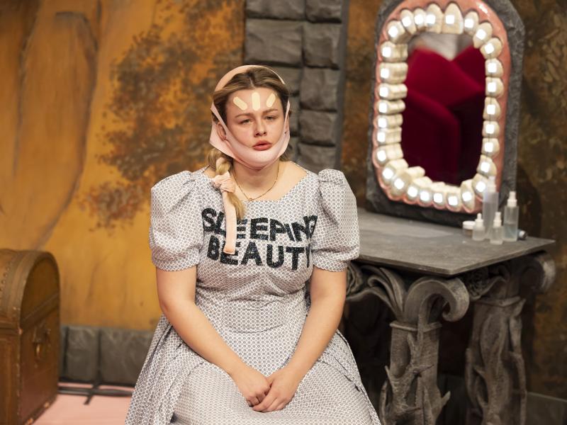 Bühnensituation: Anouk Piwek in einem hellen gepunkteten Kleid mit Puffärmeln, sitzt. Auf ihrer Brust steht „SLEEPING BEAUTY“ in schwarzen Großbuchstaben. Ihre langen blonden Haare sind eingedreht und mit rosafarbenen Bändern festgebunden. Sie trägt eine Face-Lift-Maske, auf ihre Stirn sind drei helle Pflaster geklebt. Im Hintergrund steht ein Schminktisch in Stein-Optik, um dessen Spiegel sich eine riesige Zahnreihe mit einer leuchtenden Zahnspange befindet.