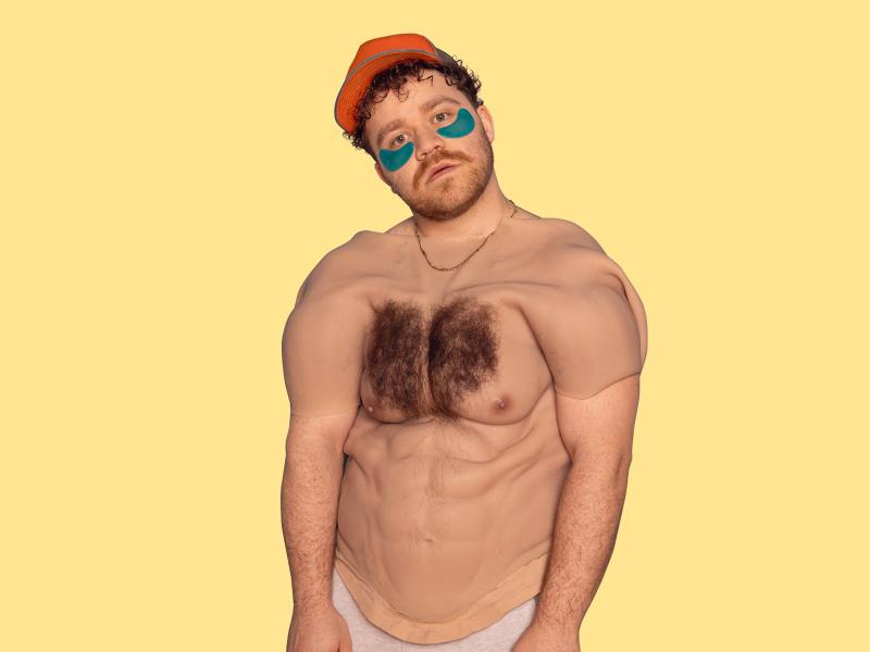 Eine männlich gelesene Person mit gelber Cap und braunen Locken und blauen Augenpads schaut in die Kamera. Über seinem Oberkörper hängt ein menschlicher Oberkörper mit behaarter Brust und Sixpack aus Silikon. Er trägt eine goldene Kette, der Hintergrund ist gelb. 