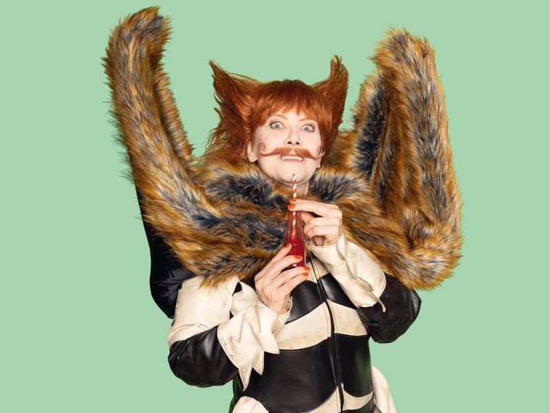 Zu sehen ist eine Person mit orangenen Haaren, die zu zwei Hörnern oder Ohren nach oben frisiert sind. Die Augen sind weit aufgerissen, sie trägt eine orangene Flasche mit einem gestreiften Strohhalm darin in den Händen. Außerdem trägt die Person einen orangenen Schnurrbart und zwei Fangzähne blitzen hervor Eine Art Fellponcho ist zu beiden Seiten nach oben geklappt. Darunter trägt die Person einen schwarz-weißen, ledrigen Anzug. Der Hintergrund ist grün.