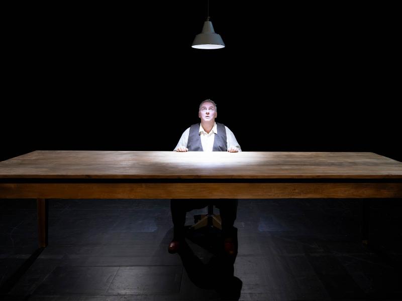 Hermann Book sitzt an einem Holztisch, über ihm leuchtet eine Hängelampe, zu der er aufblickt. Unter einer braunen Weste trägt er ein helles Hemd. Seine Hände liegen auf dem Tisch. Sein Mund ist geöffnet. Der Hintergrund und Boden sind schwarz. 
