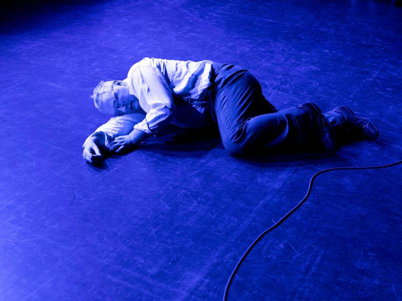 Hermann Book liegt auf dem Boden in Embryonalstellung. Alles ist in ein blaues Licht getaucht. Er trägt eine dunkle Hose und Schuhe sowie ein helles Hemd und eine Brille. Sein Mund ist leicht geöffnet und seine Augen geschlossen. 