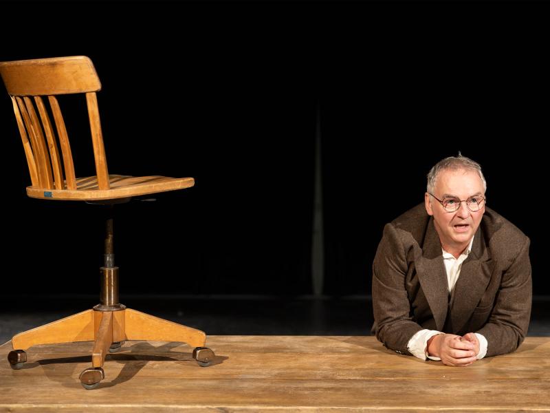 Hermann Book lehnt sich mit beiden Unterarmen auf einen Holztisch. Seine Hände sind verschränkt. Unter einem braunen Sakko trägt er ein helles Hemd, außerdem trägt er eine Brille. Beide Hände liegen auf dem Tisch. Sein Mund ist geöffnet, er schaut rechts an der Kamera vorbei. Neben ihm steht ein Holzstuhl mit Rollen auf dem Tisch. Der Hintergrund ist schwarz. 