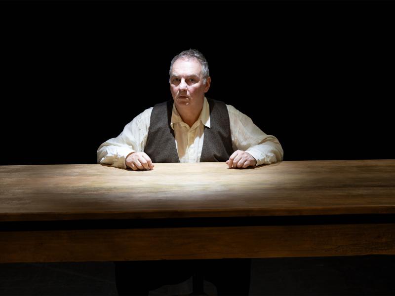 Hermann Book sitzt an einem Holztisch, nur sein Oberkörper und Kopf sind zu sehen. Unter einer braunen Weste trägt er ein helles Hemd. Beide Hände liegen auf dem Tisch. Sein Mund ist geschlossen, er schaut in die Kamera. Der Hintergrund ist schwarz. 