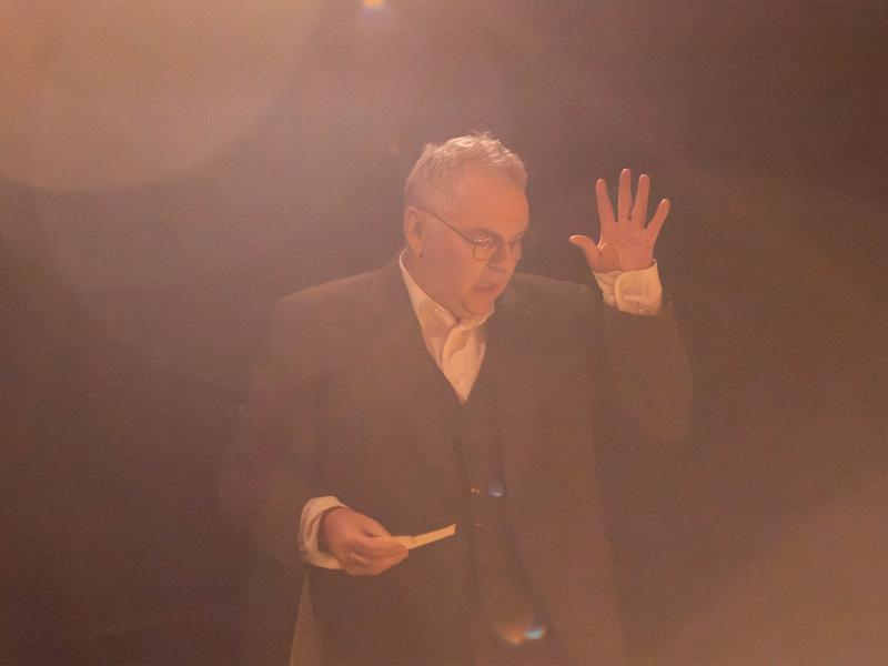 Hermann Book steht mit einem Blatt Papier in der Hand und der anderen offenen Hand erhoben vor einem schwarzen Hintergrund. Er hat den Mund geöffnet, schaut nach unten. Licht strahlt von oben und taucht die Szene in eine warme, braune Atmosphäre.  