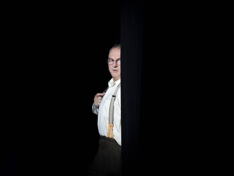 Hermann Book steht halb versteckt hinter einer schwarzen Wand, alles außer ihm ist dunkel. Er lugt hervor, trägt ein weißes Hemd, eine Brille und Hosenträger. 