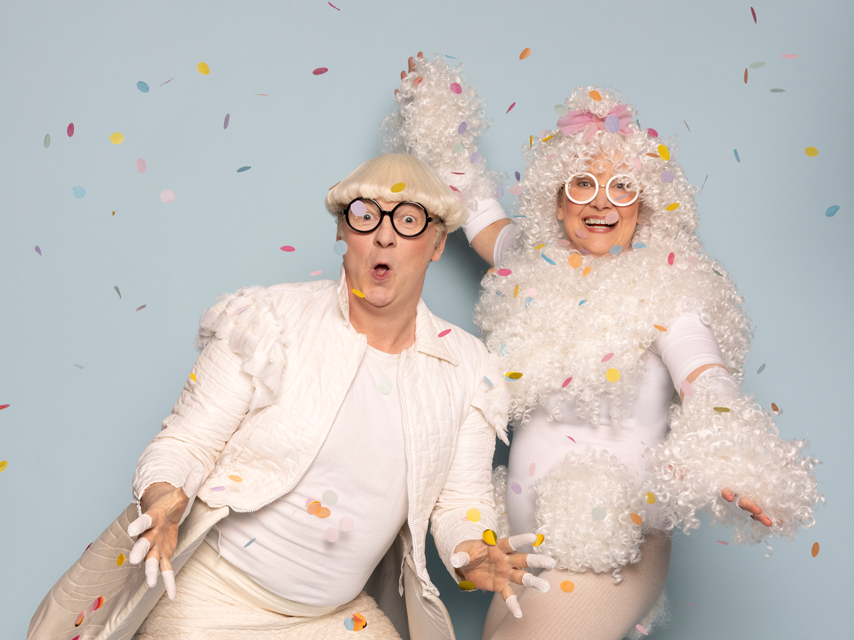 Das Bild zeigt zwei Schauspieler*innen in weißen Kostümen im Konfettiregen.