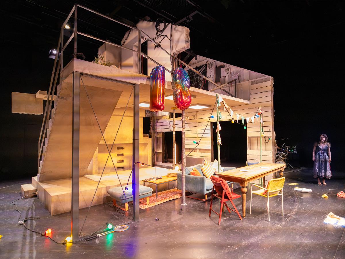 Das Bühnenbild ist verlassen und Gegenstände liegen auf dem Boden. Anastasia Lara Heller steht abseits in einem schwarzen Kleid und blickt auf die Unordnung.