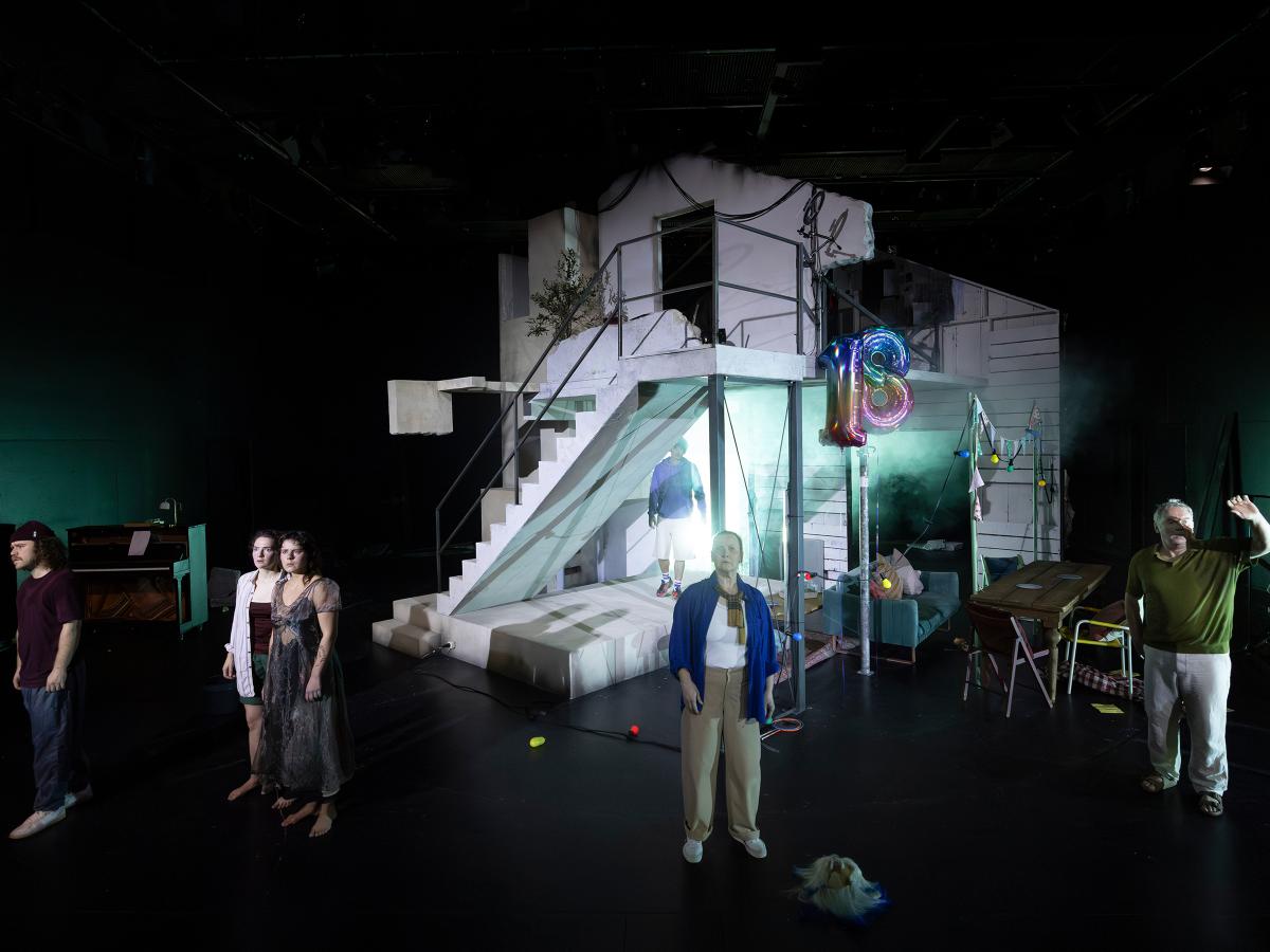 Das Ensemble steht verteilt im Bühnenbild. Alle blicken in die Ferne. Man sieht das Haus und eine Person an einem Schlagzeug im Hintergrund. 