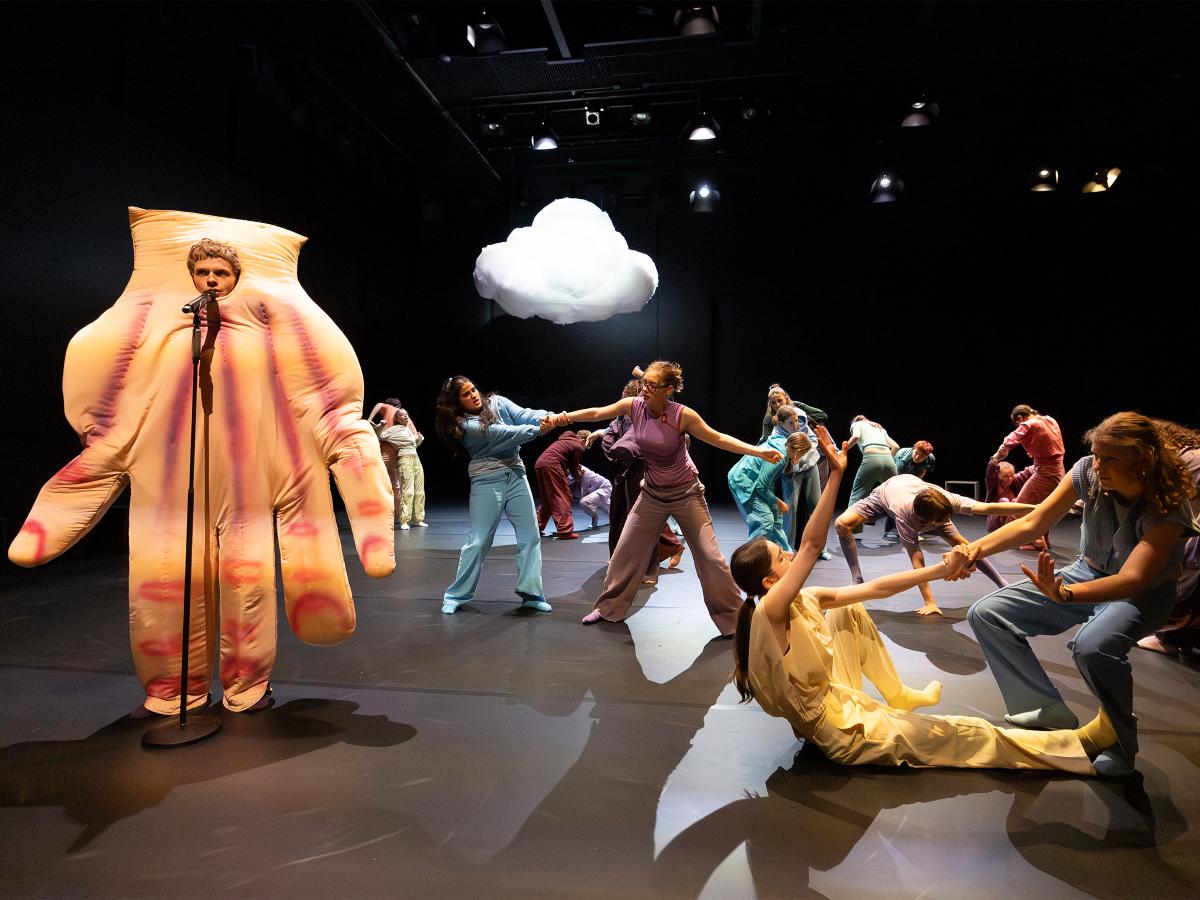 Es sind viele Tänzer*innen auf der Bühne in Bewegung. Viele tanzen in Zweierpaaren und berühren sich. Am Mikrofon steht ein Tänzer in einem Handkostüm. 