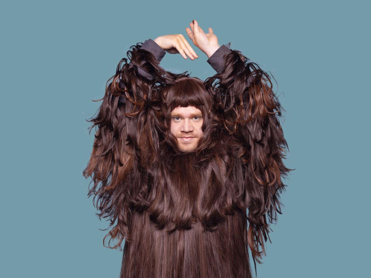 Parsa Yaghoubi Pour streckt seine Arme nach oben. Er trägt ein Kleid aus braunen Haaren, die seinen Körper, bis auf das Gesicht, komplett verdecken. Sie fallen in leichten Wellennach unten.