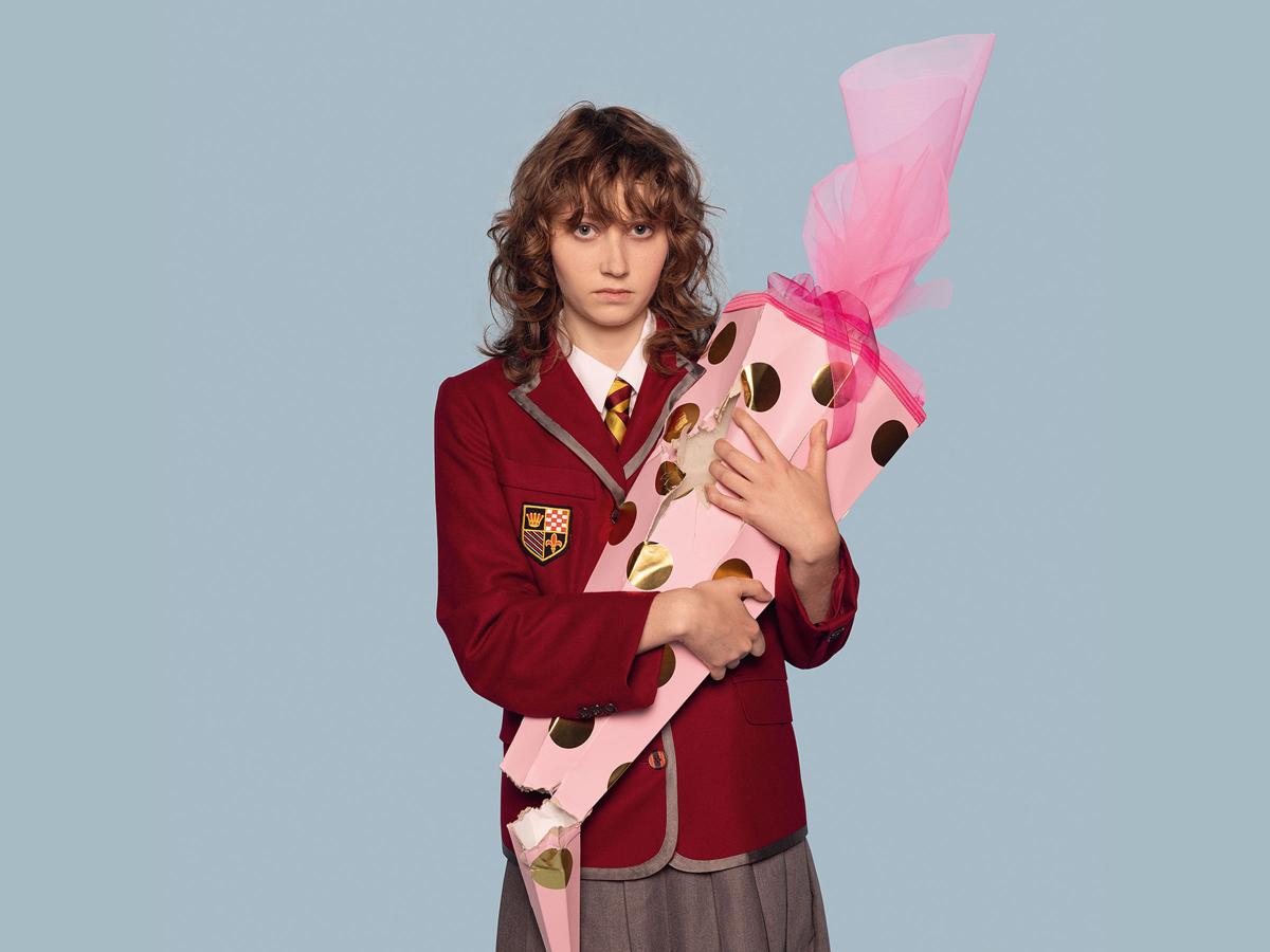 Das Bild zeigt ein jugendliches Mädchen in Schuluniform, das eine kaputte Schultüte in der Hand hält und mit sehr erschöpftem Blick in die Kamera schaut.