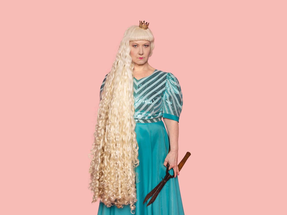 Auf dem Foto sieht man Christine Ochsenhofer in einem türkisen Kleid und einer langen, blonden Perücke. In der linken Hand hält sie eine große Schere