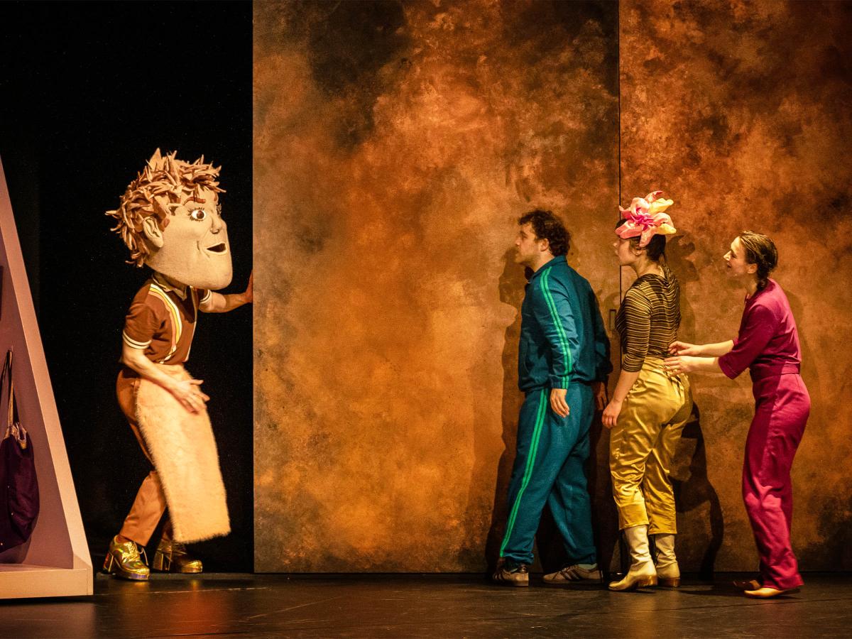 Bühnensituation: Eine Person mit braun-rosa Kleidung und einer großen Kopfmaske mit Gesicht hält sich an einer goldenen Wand fest. Sie schaut in Richtung von Parsa Yaghoubi Pour, Anastasia Lara Heller und Viktoria Kraft, die hintereinander an der gleichen Wand kurz vor der Person mit Maske stehen. 