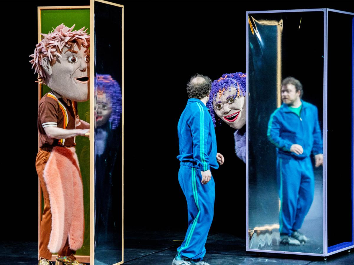 Bühnensituation: Links eine Person in braun-rosafarbener Kleidung mit einer großen Kopfmaske mit braun-rosa Haaren. Sie steht im Eingang eines hohen schmalen Kastens mit verspiegelten Wänden. Mittig steht Parsa Yaghoubi Pour in einem blauen Trainingsanzug, der sich zu einer Person mit einer weiteren Kopfmaske dreht, die lilafarbene Locken mit orangenen Strähnen und einen geöffneten, lächelnden Mund hat. Sie lehnt sich hinter einem verspiegelten hohen Kasten hervor. In diesem spiegelt sich Parsa.