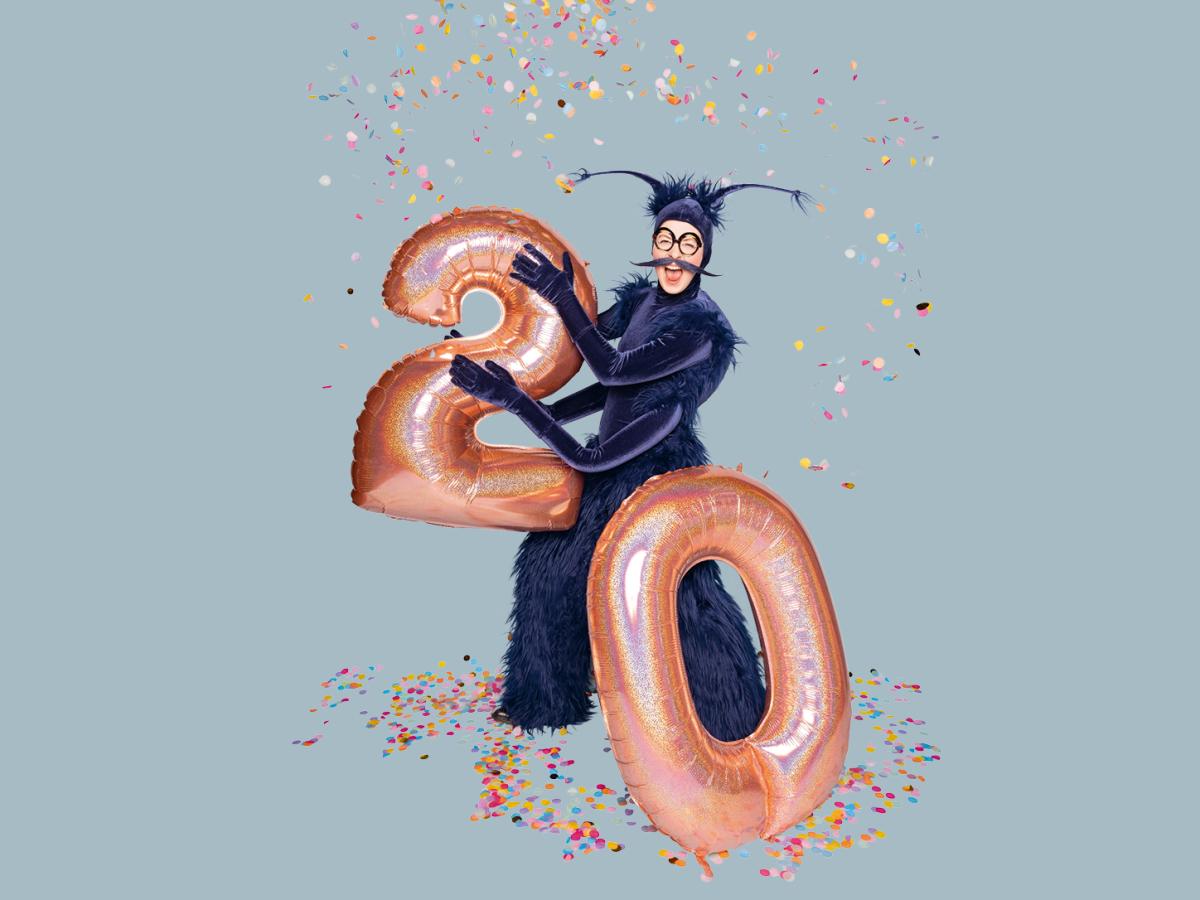 Das Foto zeigt eine Person in einem blauen Käferkostüm. Von oben fällt buntes Konfetti. In den Händen hält die Person Luftballons in Form von einer 2 und einer 0.