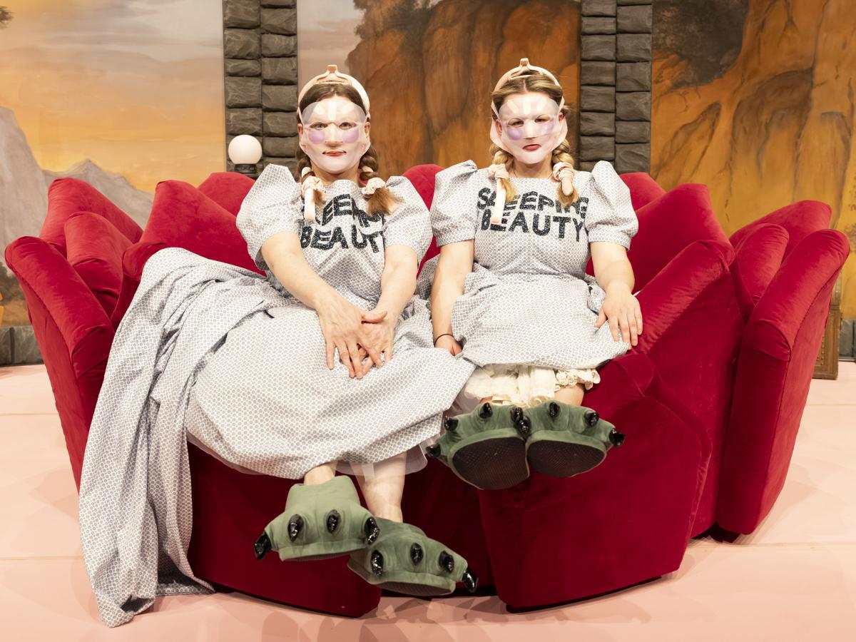 Bühnensituation: Christine Ochsenhofer und Anouk Piwek, beide in einem hellen gepunkteten und Kleid mit Puffärmeln sitzen auf einem roten Plüschsofa mit Zacken, die Hände in den Schoß gelegt. Auf ihrer Brust steht „SLEEPING BEAUTY“ in schwarzen Großbuchstaben. Ihre langen blonden Haare sind eingedreht und mit rosafarbenen Bändern festgebunden. Sie tragen Face-Lift-Masken, die Gesicht bis auf Mund und Augen vollständig bedecken. An ihren Füßen befinden sich große grüne Puschen mit schwarzen Krallen daran.