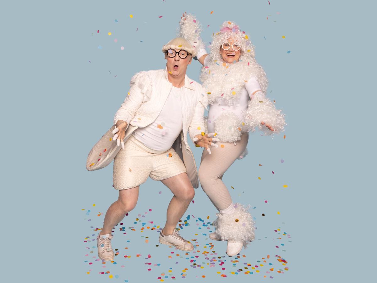 Das Bild zeigt die Schauspieler*innen Herman Book und Christine Ochsenhofer in weißen Kostümen inmitten von Konfetti