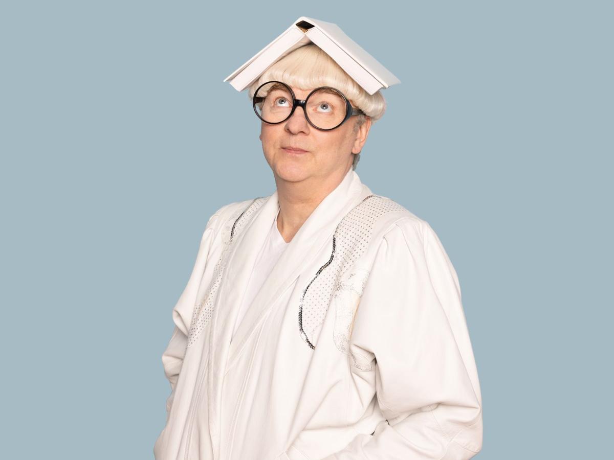 Das Bild zeigt den Schauspieler Hermann Book in weißem Kostüm, mit weißer Perücke und großer, runder Brille. Auf seinem Kopf liegt ein aufgeklapptes Buch.