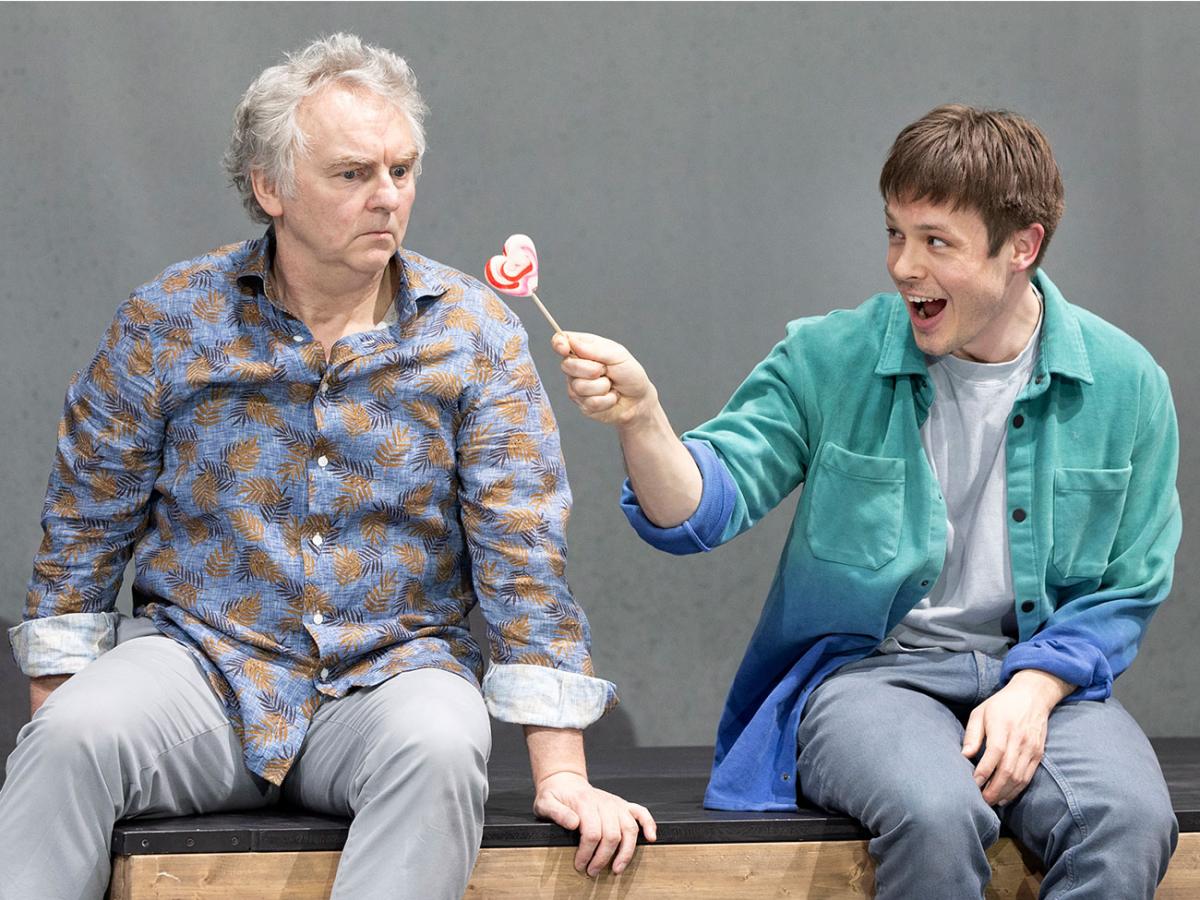 Das Bild zeigt den Schauspieler Hermann Book, der grimmig schaut. Der Schauspieler Severin Mauchle sitzt zu seiner Rechten und hält ihm freudig einen Lolli in Herzform hin.