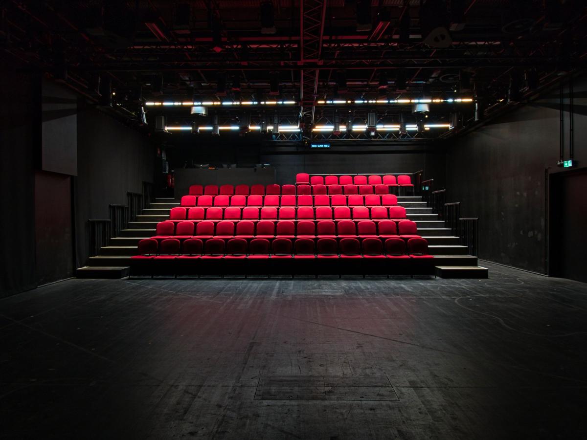 Zu sehen ist die Studiobühne im Jungen Schauspielhaus. Es handelt sich um einen schwarz gehaltenen Bühnenraum mit einer Beleuchtungseinrichtung und einer Zuschauertribüne mit roten Sitzen.