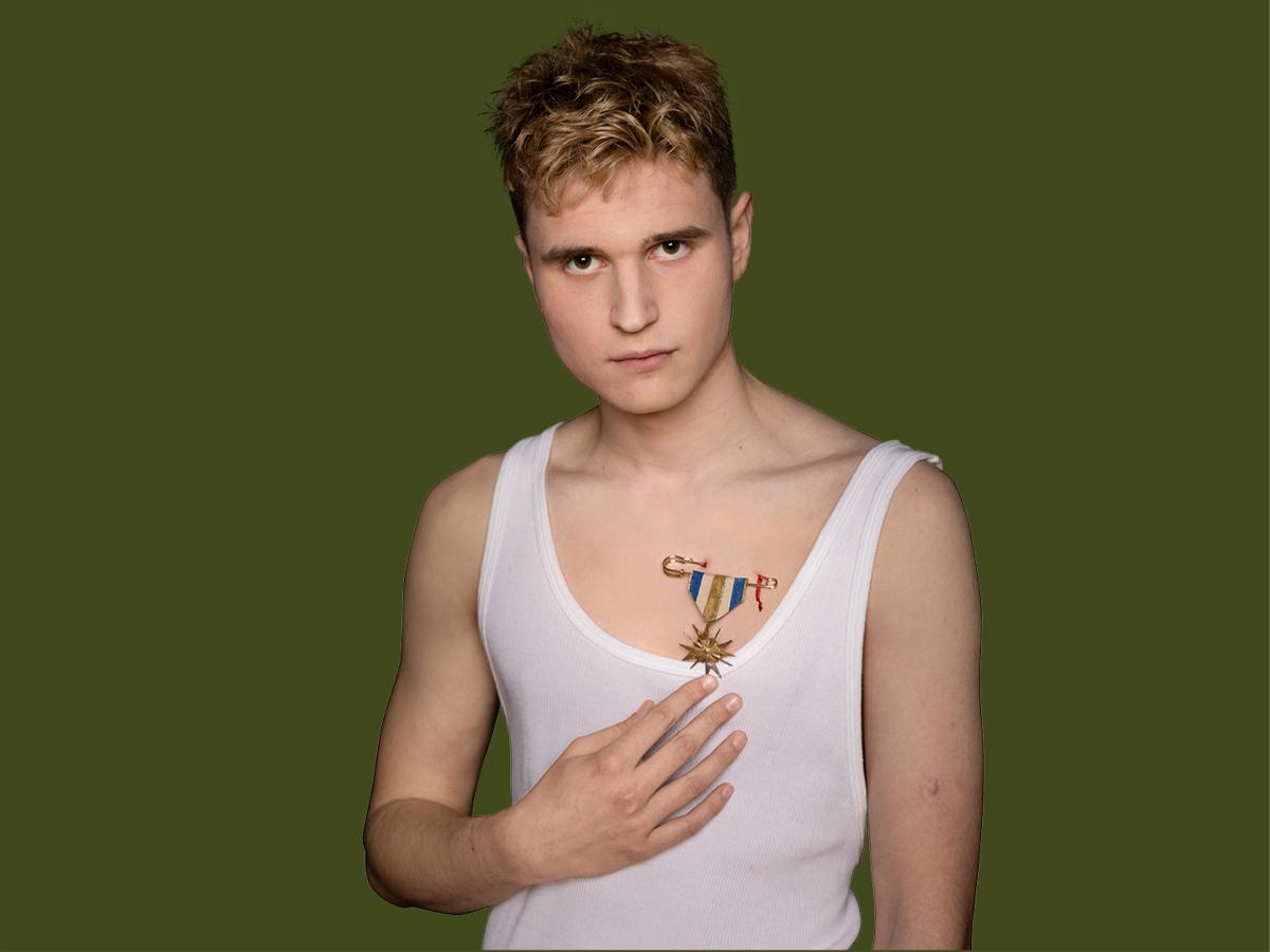 Eine männlich gelesene Person mit blonden kurzen Locken in weißem Tanktop ist zu sehen. Eine Hand deutet auf eine Brosche, die an ein Abzeichen erinnert und in die Haut sticht. Die Person schaut in die Kamera. Der Hintergrund ist dunkelgrün.