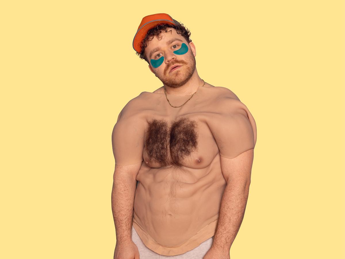 Eine männlich gelesene Person mit gelber Cap und braunen Locken und blauen Augenpads schaut in die Kamera. Über seinem Oberkörper hängt ein menschlicher Oberkörper mit behaarter Brust und Sixpack aus Silikon. Er trägt eine goldene Kette, der Hintergrund ist gelb. 