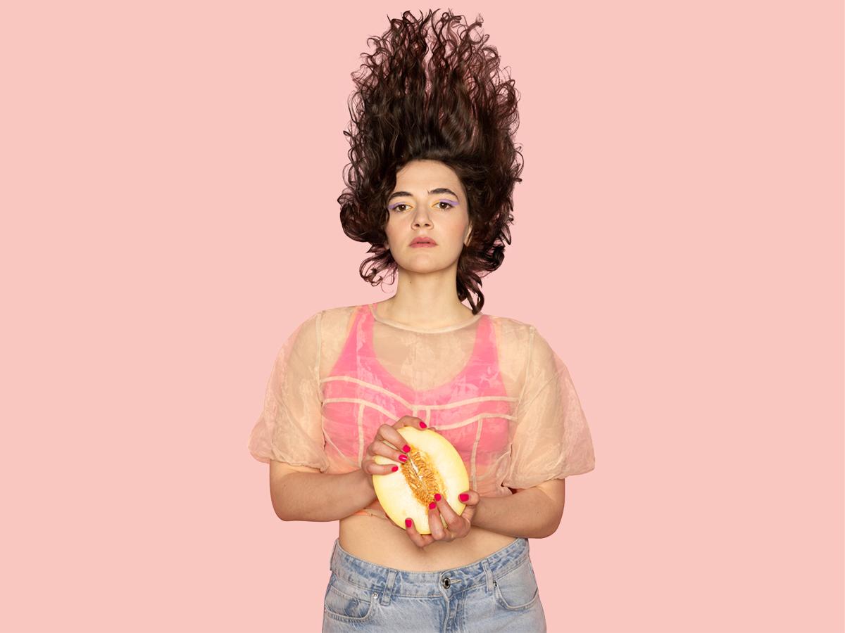 Eine weiblich gelesene Person hat braune Locken in die Luft geworfen. Sie trägt einen pinker Sport-BH, darüber ein durchsichtiges T-Shirt und eine Jeans. Ihre pinken Fingernägel greifen in eine aufgeschnittene Melone. Der Hintergrund ist pink.