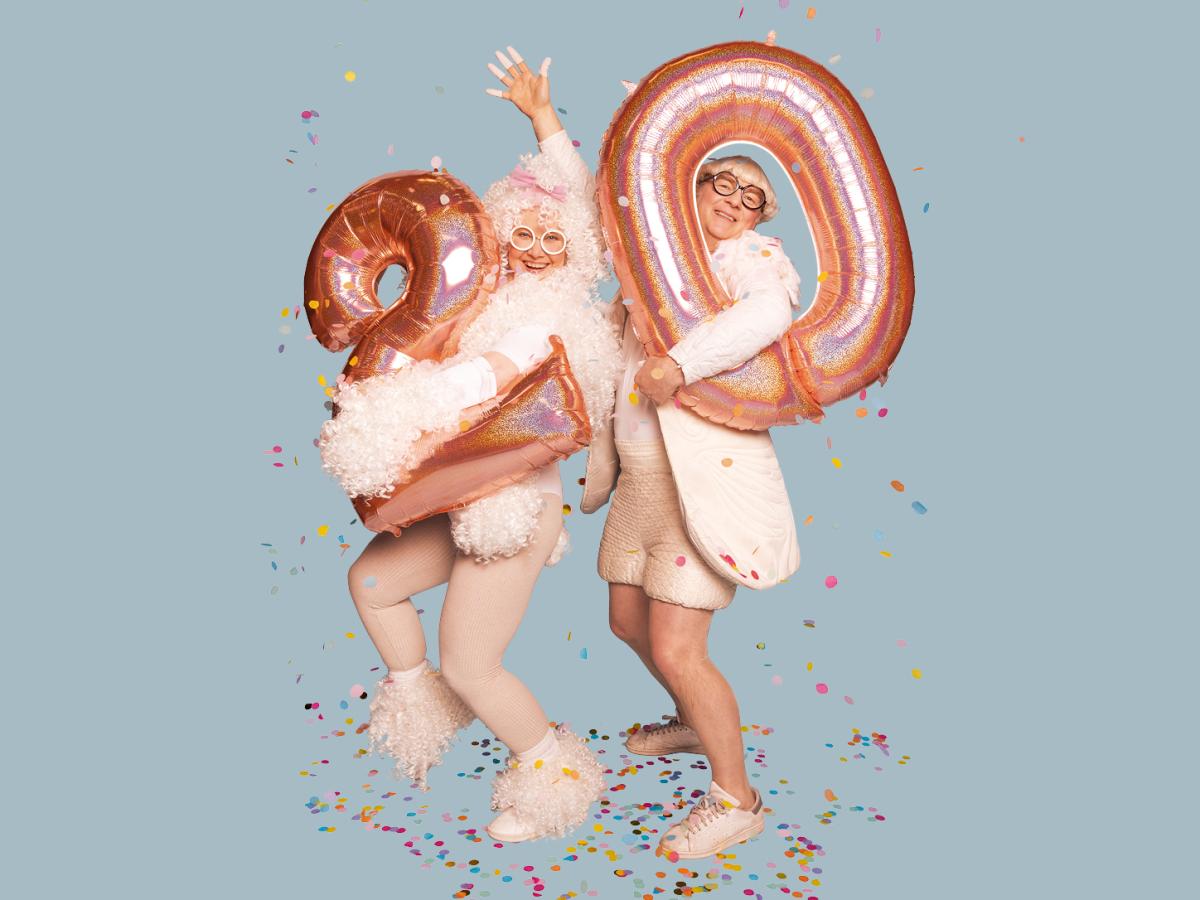 Das Bild zeigt die beiden Schauspieler*innen Hermann Book und Christine Ochsenhofer. Sie stehen im Konfettiregen und halten Luftballons mit den Zahlen 2 und 0 in den Händen.