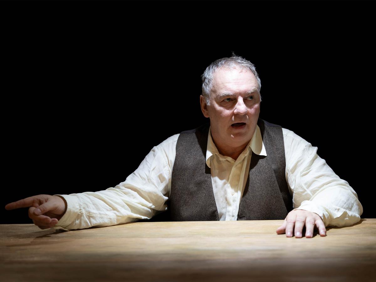 Hermann Book sitzt an einem Holztisch, nur sein Oberkörper und Kopf sind zu sehen. Unter einer braunen Weste trägt er ein helles Hemd. Eine Hand liegt auf dem Tisch, mit der anderen zeigt er nach links. Sein Mund ist geöffnet, er schaut nach rechts. Der Hintergrund ist schwarz. 